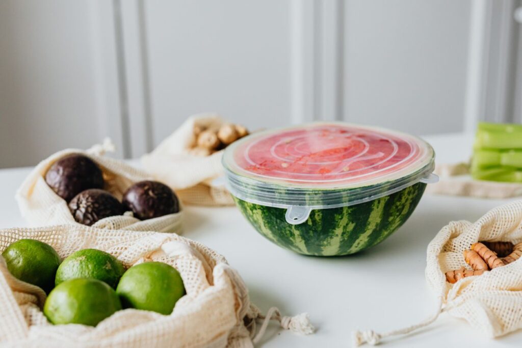 A fresh sliced watermelon with reusable food wrap, surrounded by avocados and limes on a white table